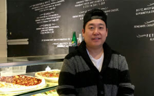 Portrait of the Lean Crust pizza shop owner wearing a black and white striped sweater and a black baseball cap on backwards with pizzas behind him in a case and black wall with white writing describing the menu.