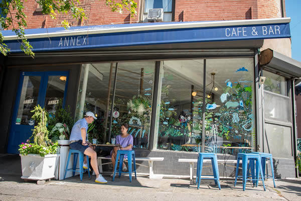 An exterior view of the Greene Grape annex with a blue awning that reads Annex Cafe and Bar in white with blue painted metal stools with two people sitting at an outdoor table having a drink.