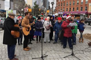 People in winter gear, some with Santa hats, gathered around microphones in Fowler Plaza singing holiday music with one man playing the guitar.