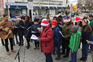 People in winter gear, some with Santa hats, gathered around microphones in Fowler Plaza singing holiday music with one man playing the guitar.