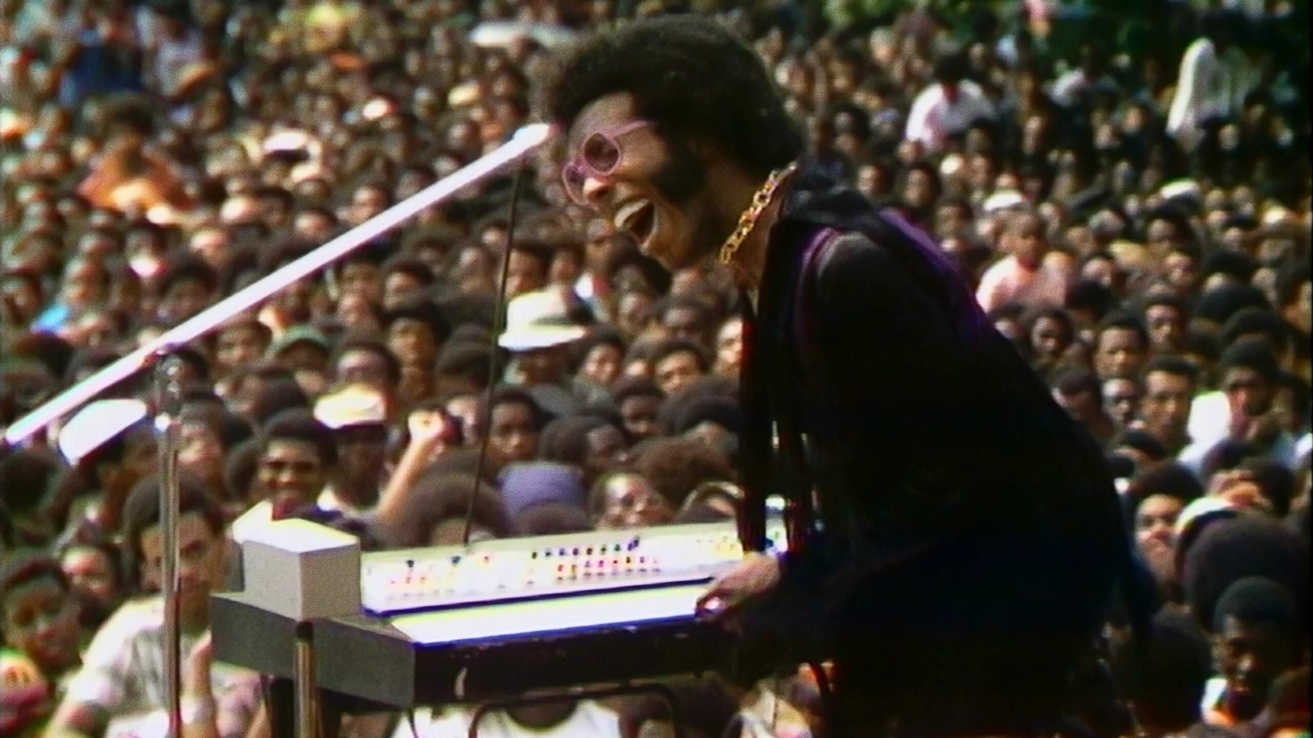 Image is archive photo of Sly and The Family Stone on stage, with concert crowd in background