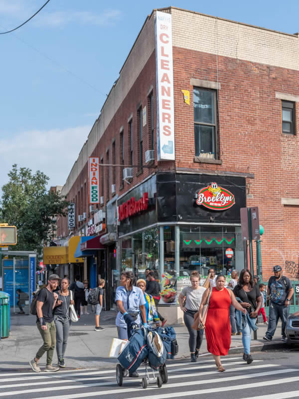 A group of people crossing the street at a Brooklyn intersection on a summer day showing several businesses in the background including a dry cleaner and an Indian restaurant.