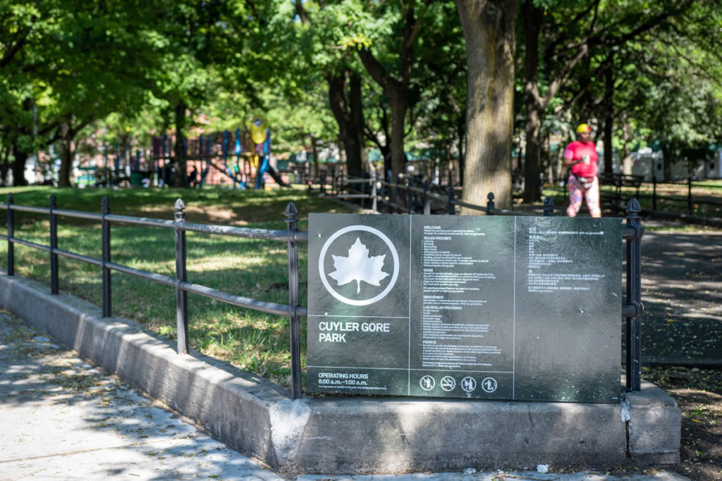 A green sign with a giant white maple leaf in a circle over the name Cuyler Gore Park on a black iron fence in the foreground with a park and person walking in the background on a summer day.