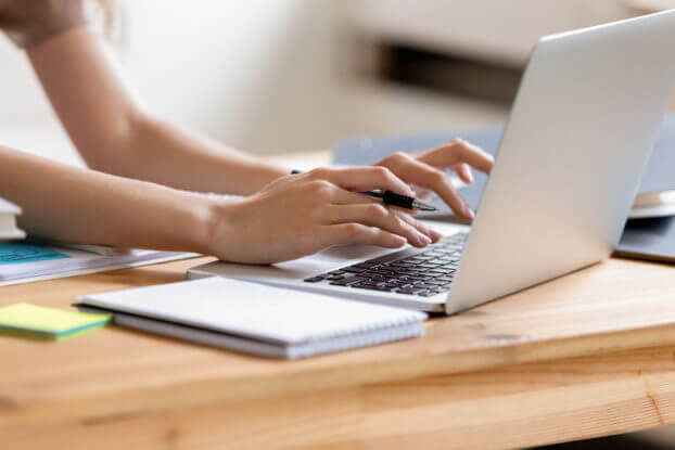 A scene of a woman typing on laptop with a writing pad nearby.